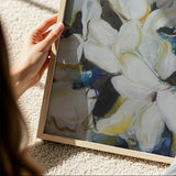 Hand holding a framed abstract painting with white flowers on a textured surface