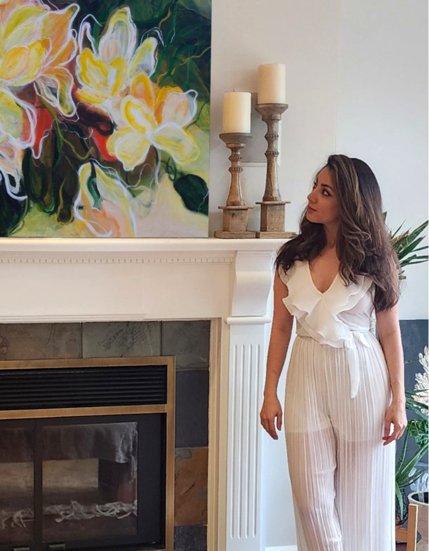 Mona, the artist standing next to one of her original paintings on a fireplace. The painting is a 48 x 48 Florecer abstract original in the colours of green oranges, yellow, beige and white.
